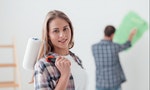 Home renovation, decoration and painting: young woman posing with a paint roller, her boyfriend is working on the background