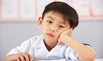 Unhappy Male Student Working At Desk In Chinese School Classroom