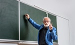 Senior lecturer writing something on blackboard and showing one finger up — Photo by IgorVetushko