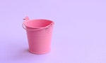 A miniature empty pink bucket lies on a violet pastel background. Minimal concept