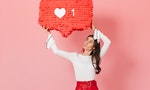 Portrait of girl with interest looking at huge ""like"" sign from Instagram. Brunette in red skirt posing on pink background