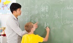 friendly elementary school teacher helping young boy writing chinese on chalkboard