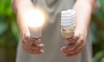 Power saving concept. Hands holding new Light Emitting Diode ( LED ) light bulb with light on and blur spiral compact-fluorescent (CFL) bulbs behind for copyspace.