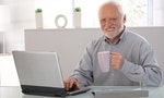 Mature man using computer, drinking tea, looking at camera, smiling.